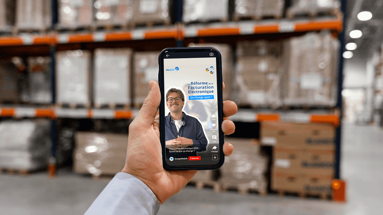 Main d'un travailleur dans un entrepôt, qui nous montre son téléphone. L'écran montre la vidéo de WeSoft sur la réforme de la facturation électronique